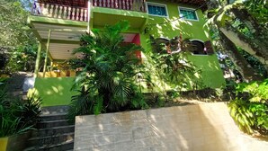 Un edificio a due piani con facciata verde e tetto in tegole rosse. Una scala conduce all'ingresso, circondata da un'abbondante vegetazione.