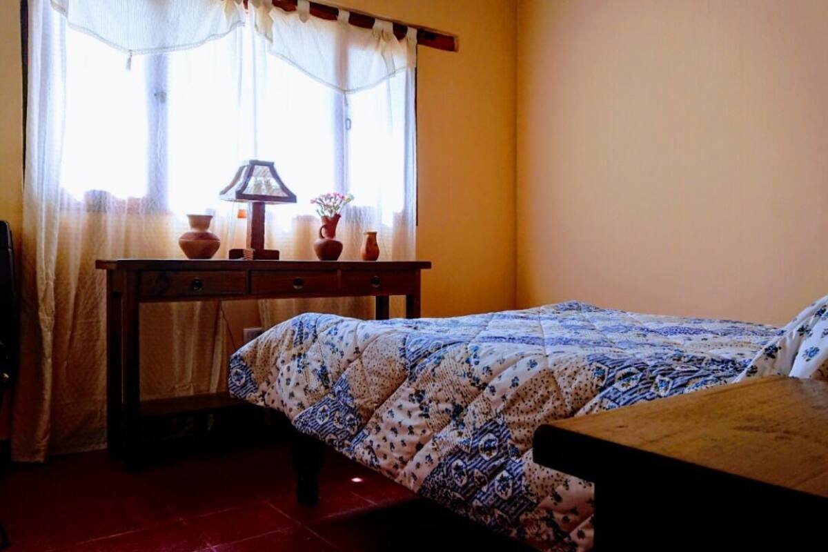 A bedroom with a bed, a wooden nightstand, a desk with a lamp, and a window with curtains.