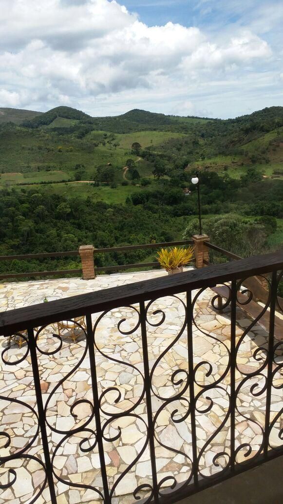 A balcony with a stone floor and metal railing, overlooking a green landscape with hills and a clear sky.