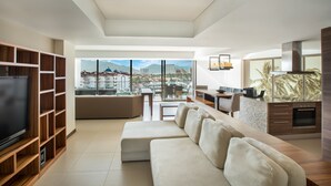 A modern living room with a large flat-screen TV, a beige sectional sofa, wooden shelving, and a dining area with a view of the city and mountains. - Nima Bay Lifestyles Experience (Puerto Vallarta)