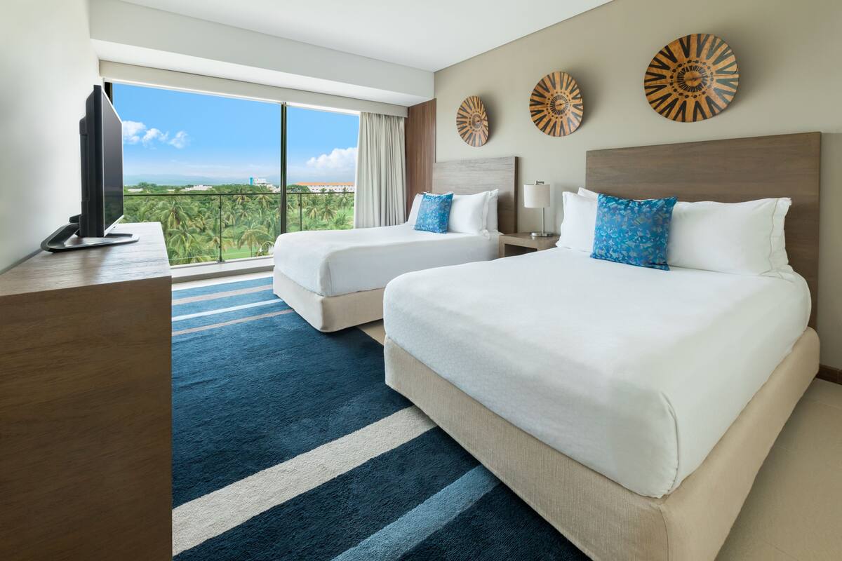 Een hotelkamer met twee bedden, een televisie en een groot raam met uitzicht op palmbomen.