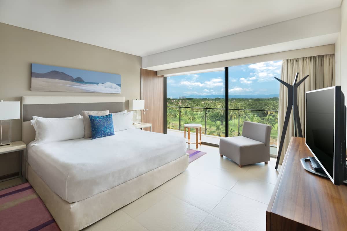 Een moderne hotelkamer met een groot bed, een televisie, een bureau en een balkon met uitzicht op groen.