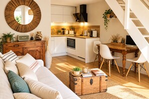 A compact living space with a sofa, a wooden chest, a dining table with chairs, a kitchen area, and a staircase.
