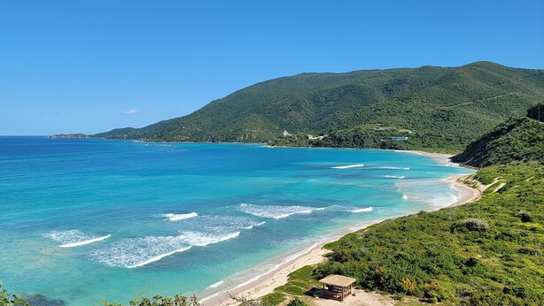 Un paisaje costero con aguas turquesas cristalinas, una playa arenosa y colinas verdes exuberantes al fondo.