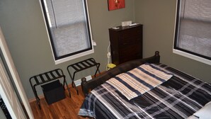 Een slaapkamer met een bed, een dressoir, een stoel en twee ramen met jaloezieën.