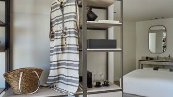 A modern hotel room with a bed, a wardrobe, a vanity with a mirror, and a shelf with a basket and a towel.