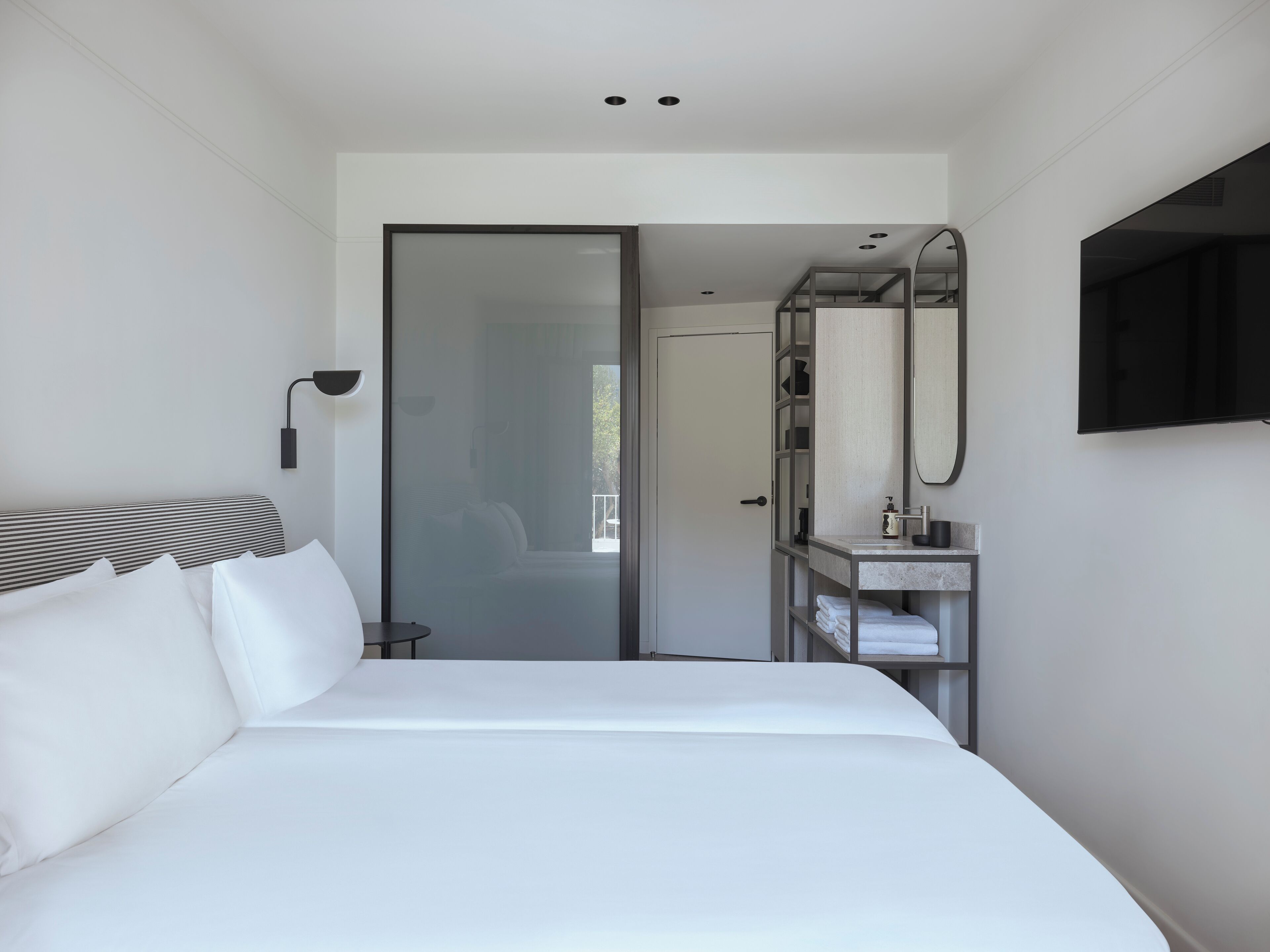 A modern hotel room with a large bed, a bedside table, a mirror, and a bathroom visible through a glass door.