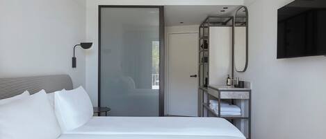 A modern hotel room with a large bed, a bedside table, a mirror, and a bathroom visible through a glass door.