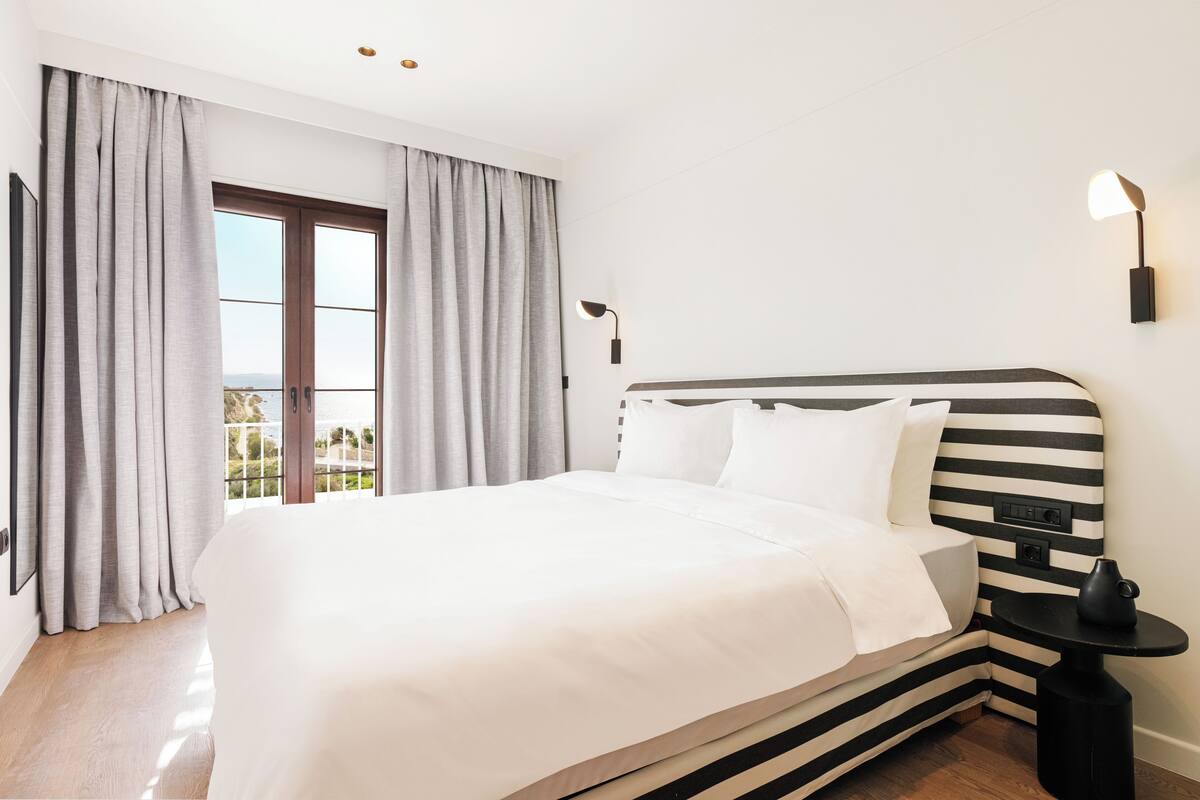 A modern hotel room with a large bed, grey curtains, a black and white striped headboard, a small side table, and a wall-mounted lamp.