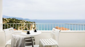 A balcony with white chairs and a small table, overlooking a coastal landscape with a view of the sea and distant mountains.