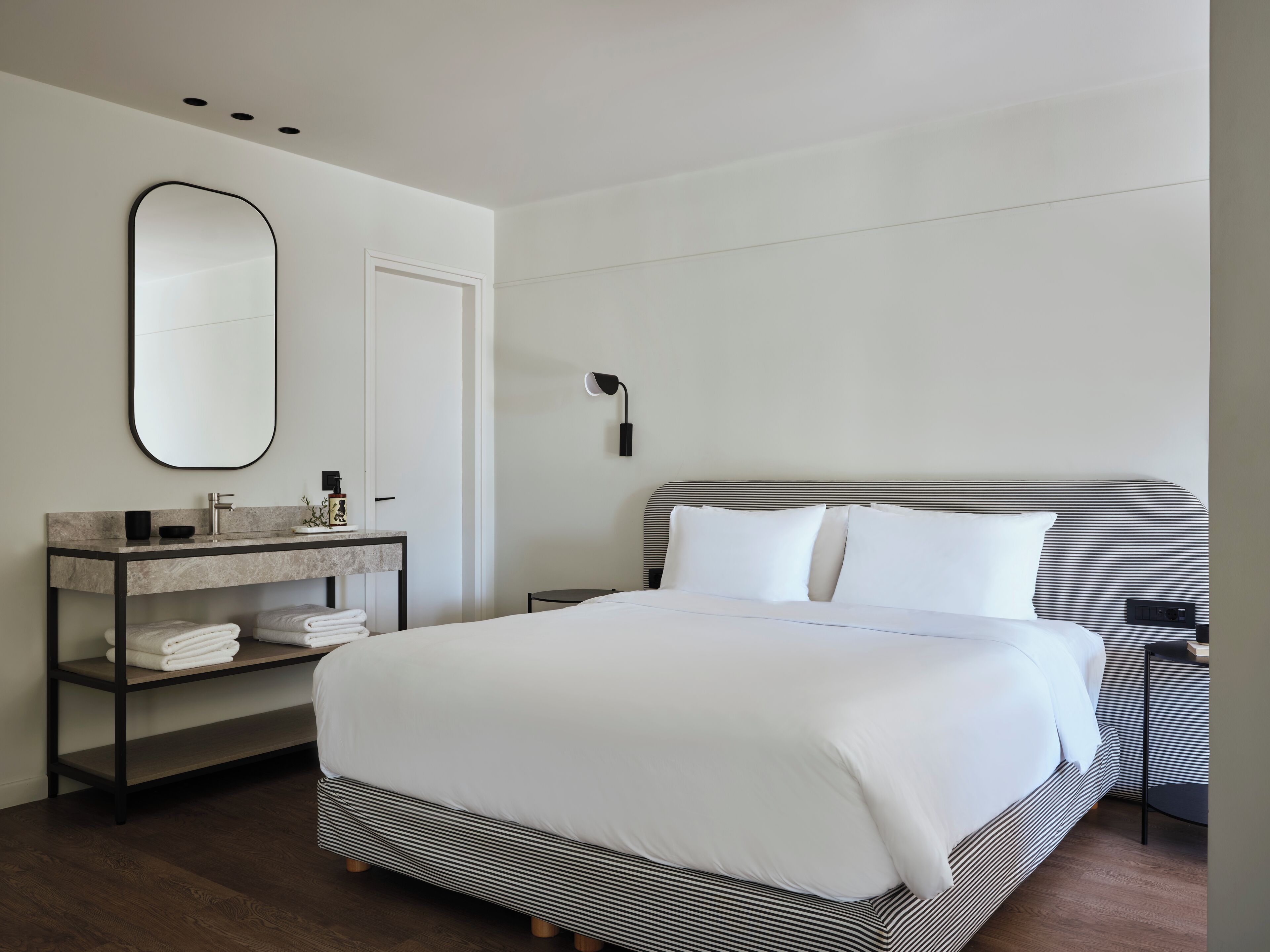 Une chambre d’hôtel moderne avec un grand lit, une table de chevet, un miroir et une petite étagère avec des serviettes.