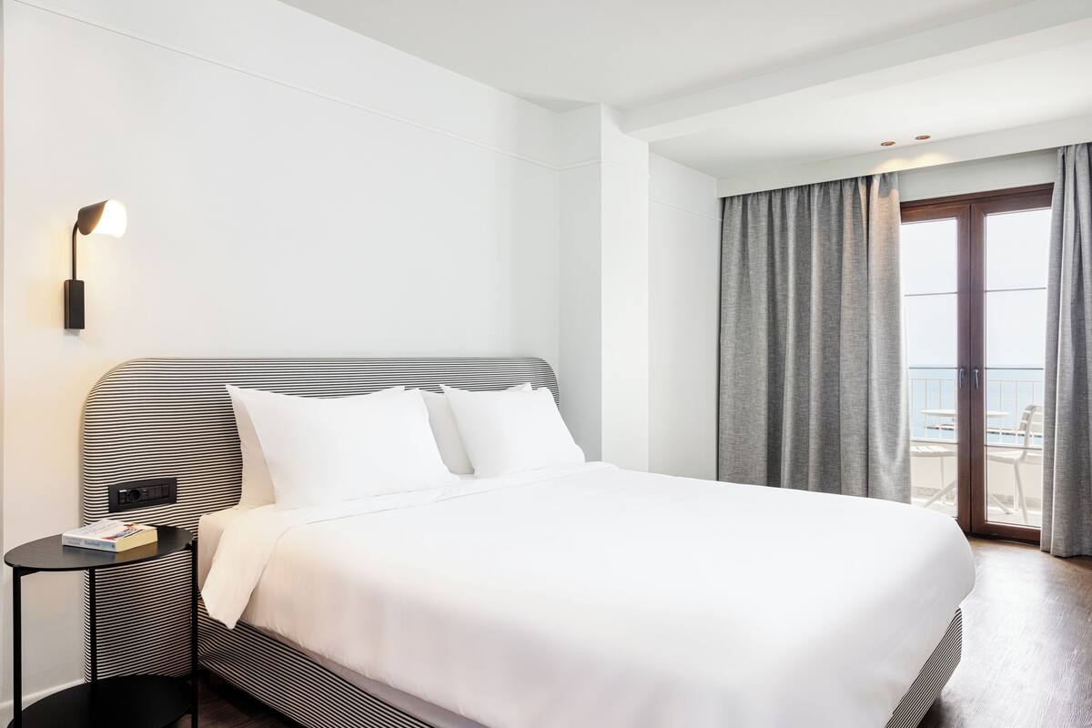 A modern hotel room with a large bed, a bedside table, a lamp, and a view of the outdoors through a sliding door.