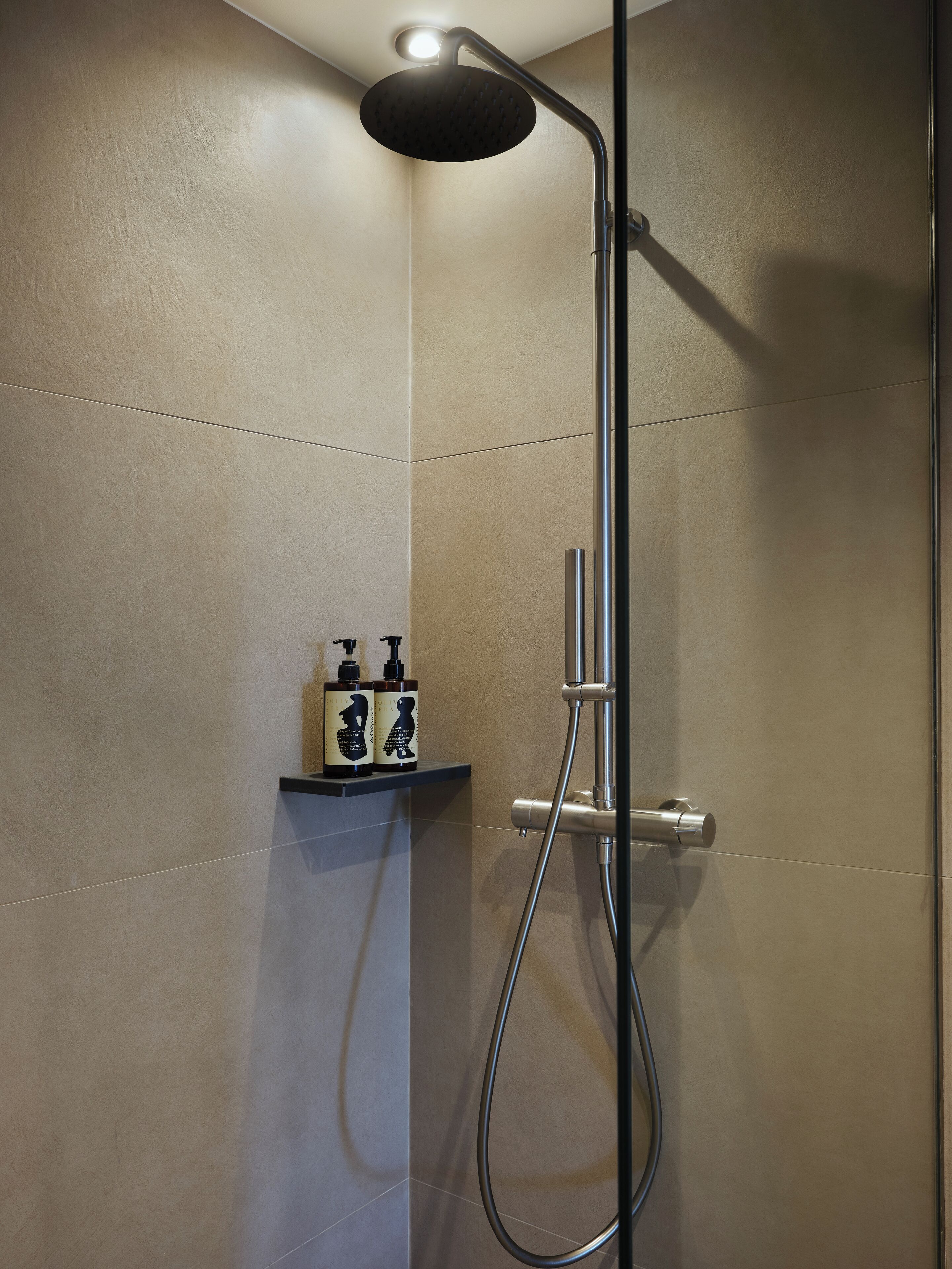 A modern shower with a handheld showerhead, a handheld shower attachment, and a wall-mounted faucet. There is a shelf with two bottles of shampoo and conditioner.