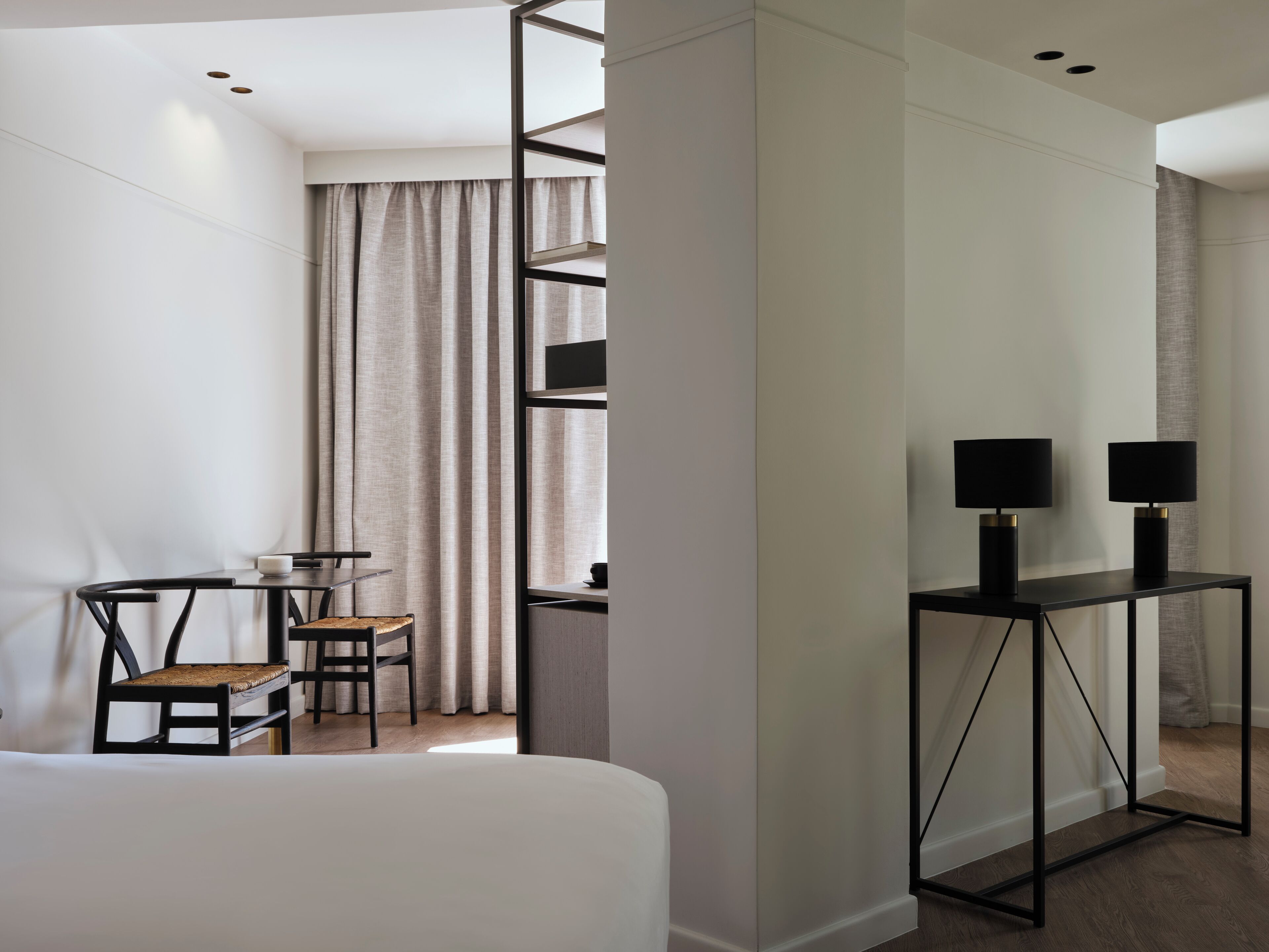 A hotel room with a bed, a desk with two lamps, a chair, and a window with curtains.