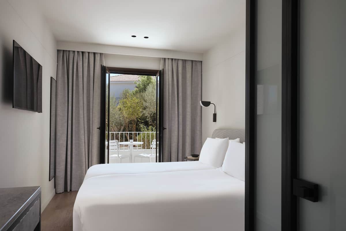 A modern hotel room with a large bed, a balcony with a table and chairs, and a view of greenery outside.
