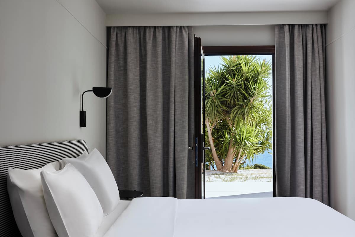 A modern hotel room with a large bed, grey curtains, a side table, and a view of a palm tree and beach.