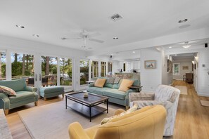 A spacious living room with a large window, a ceiling fan, and a kitchen area visible through an open door. - Botanical Garden Escape on the River - with a Pool (Lancaster)