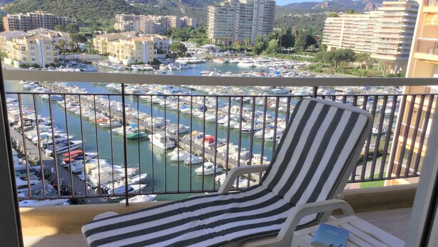 A balcony with a striped chair and a small table, overlooking a marina with boats and buildings in the background. - Apartment "France II" overlooking Cannes Marina and the Lerins Islands (Mandelieu-la-Napoule)