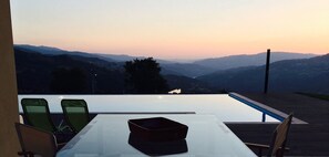 Uma área de refeições com mesa e cadeiras, com vista para uma paisagem montanhosa ao pôr do sol.