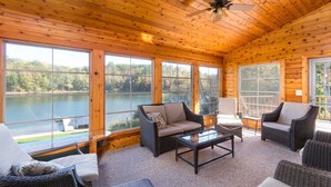 Une cabane en bois dotée de grandes fenêtres donnant sur un lac, meublée avec des meubles en osier et équipée d’un ventilateur de plafond.