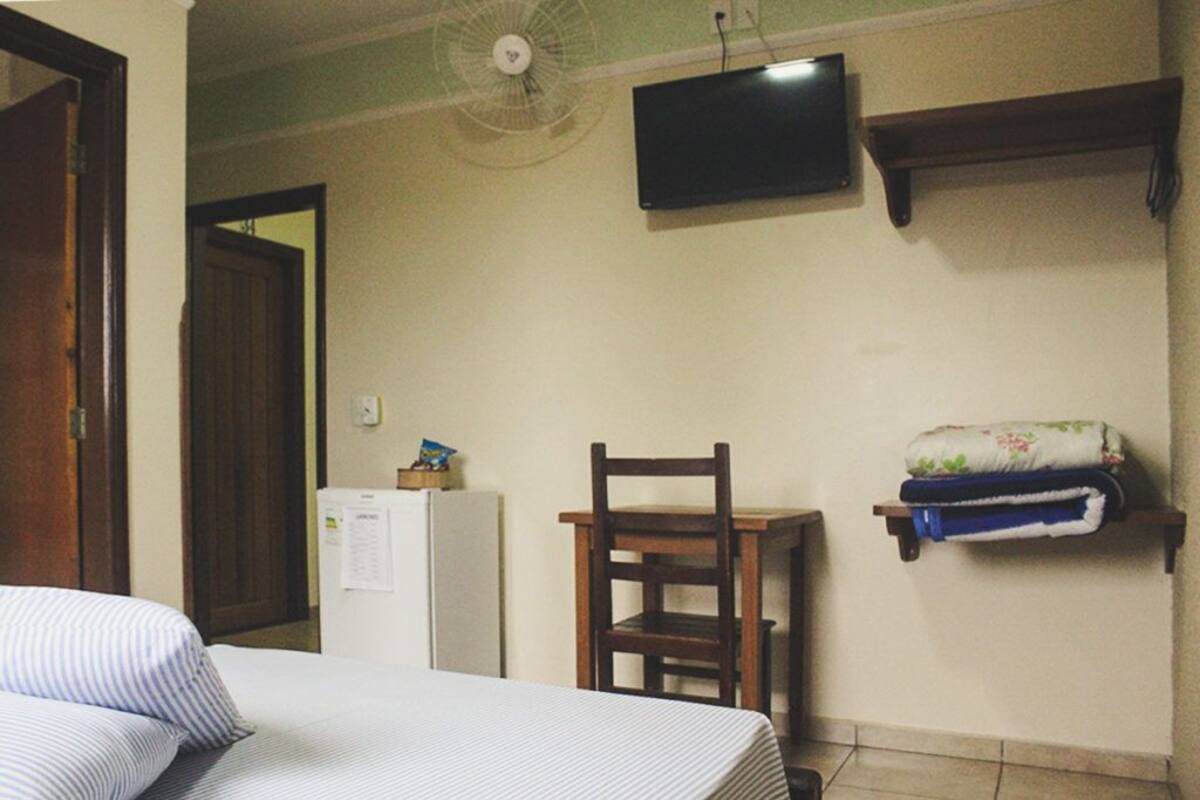 A hotel room with a bed, a chair, a television, a fan, a refrigerator, and a shelf with towels.