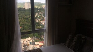 A bedroom with a large window, a bed with a leopard print bedspread, and a view of a cityscape with buildings and greenery. - 27th Flr Condo 2 w/WIFI @ IT Park Avida Riala Twrs (Cebu City)
