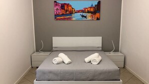 Una cama bien hecha con dos toallas enrolladas, un cabecero gris y un cuadro de una escena de canal encima.