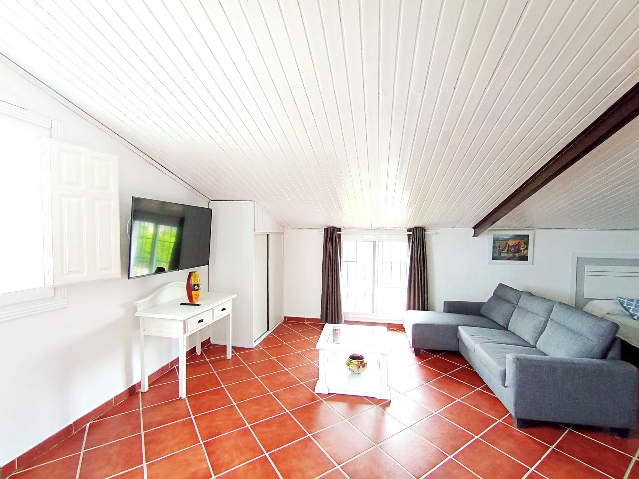 Una sala de estar con un escritorio blanco, un sofá gris, un televisor montado en la pared y una ventana con cortinas.