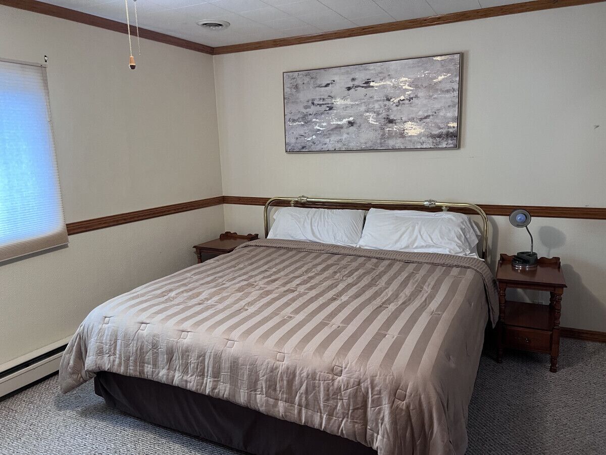 A bedroom with a large bed, a bedside table with a lamp, a window with blinds, and a painting on the wall.