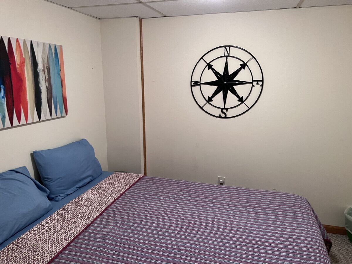 A bedroom with a bed, blue pillows, a patterned bedspread, a compass wall decoration, and a colorful abstract painting.