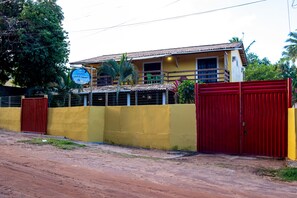 A yellow building with a red gate, a sign indicating a residential area, and a fenced yard. - Pousada Pêlo De Imbassai (Mata de São João)