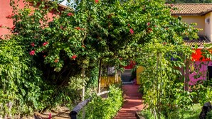 A pathway lined with lush greenery and flowering plants, leading to a building with a red roof. - Pousada Pêlo De Imbassai (Mata de São João)