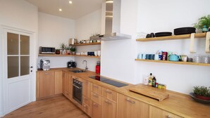 Eine kompakte Küche mit Holzausstattung, einem Backofen, einer Mikrowelle und einem Spülbecken. Über der Arbeitsplatte befinden sich Regale mit Geschirr und einem Wasserkocher.