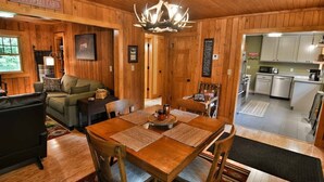 Interior de una cabaña con mesa de comedor, sillas, sofá, chimenea y zona de cocina.