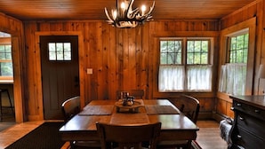 Un interior de madera con una mesa de comedor preparada para cuatro, una araña y una ventana con cortinas.