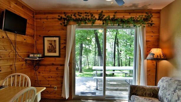 L’intérieur d’un chalet en bois comprend une table à manger, des chaises blanches, un canapé, un téléviseur fixé au mur et une grande porte-fenêtre coulissante qui donne sur une terrasse extérieure.