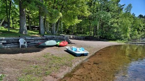 Un terrain de camping au bord d’un lac, avec une étendue d’eau claire, une chaise blanche, ainsi que plusieurs kayaks et un canoë.