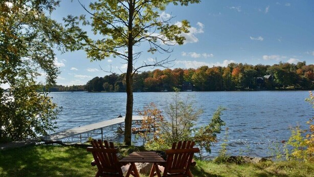 Een zitgedeelte aan het meer met twee Adirondack-stoelen en een houten tafel, met uitzicht op een rustig meer met een steiger en herfstkleurige bomen op de achtergrond.