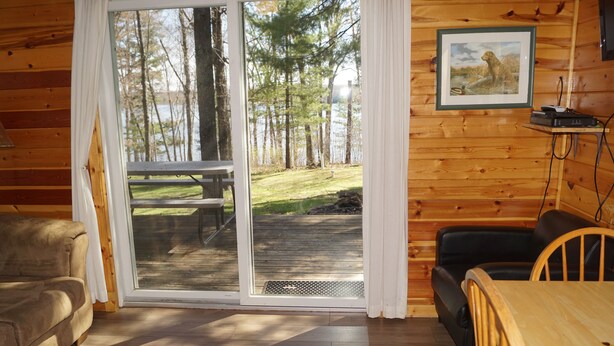 Interior de una cabaña con piso de madera, un sofá, una mesa de comedor y un ventanal corredizo que da a una terraza con vistas a árboles y un lago.