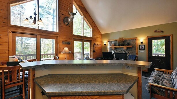 A cabin interior with a kitchen, dining area, and a large window overlooking a forest. - Deer Trail Log Home #13  on Lake Delta near Iron River, WI (Iron River)