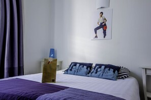 Een net opgemaakt bed met een paars dekbed en twee kussens met een abstract blauw ontwerp. Boven het bed hangt een poster van een persoon in vrijetijdskleding.