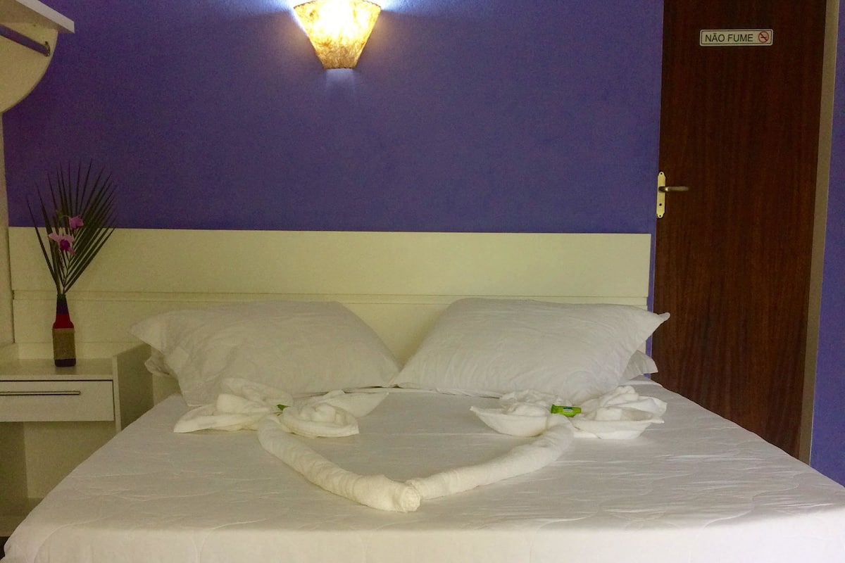 A neatly made bed with a towel arrangement, a nightstand with a vase of flowers, and a wall-mounted light fixture.