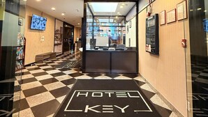 Un vestíbulo de hotel con suelo a cuadros que lleva a la recepción, un televisor montado en la pared y diplomas enmarcados en la pared.