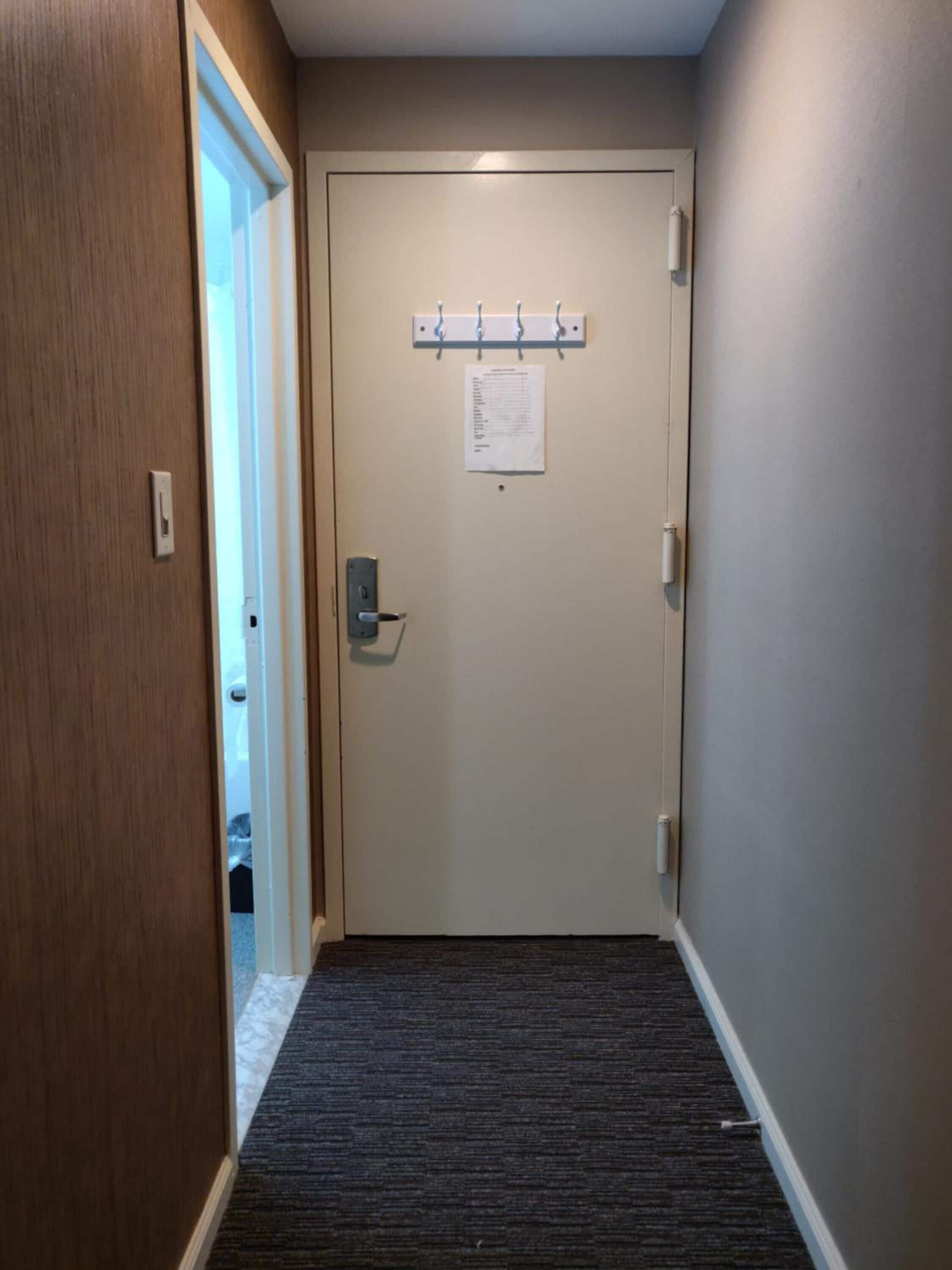 Um corredor com piso coberto por carpete, uma porta com interruptor de luz e um porta-roupas fixado na parede com ganchos.
