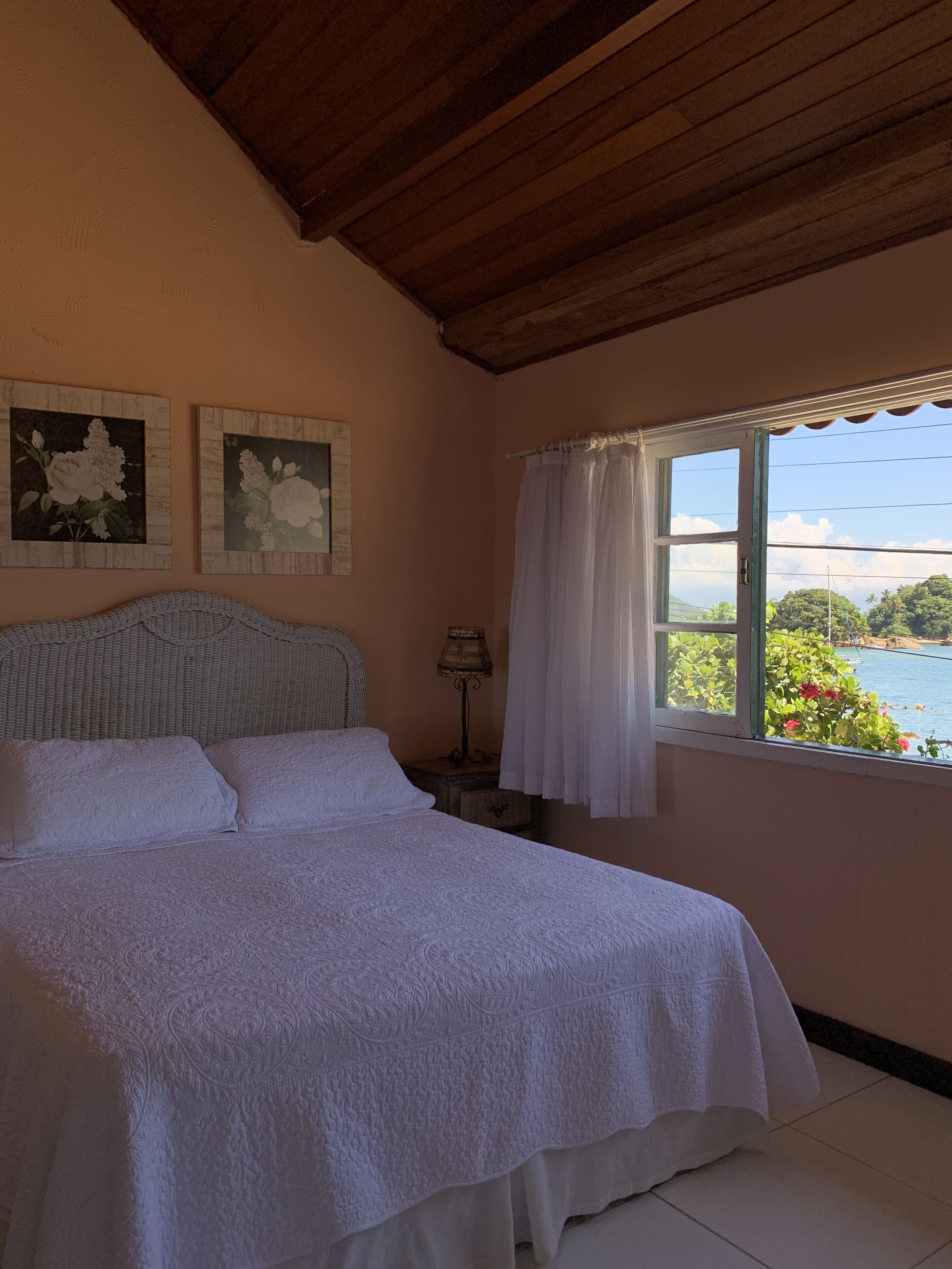 A bedroom with a bed, a nightstand, a lamp, and a window with a view of greenery and water.