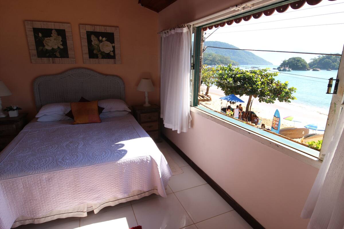 A bedroom with a bed, a nightstand, a window with a view of a beach and boats.