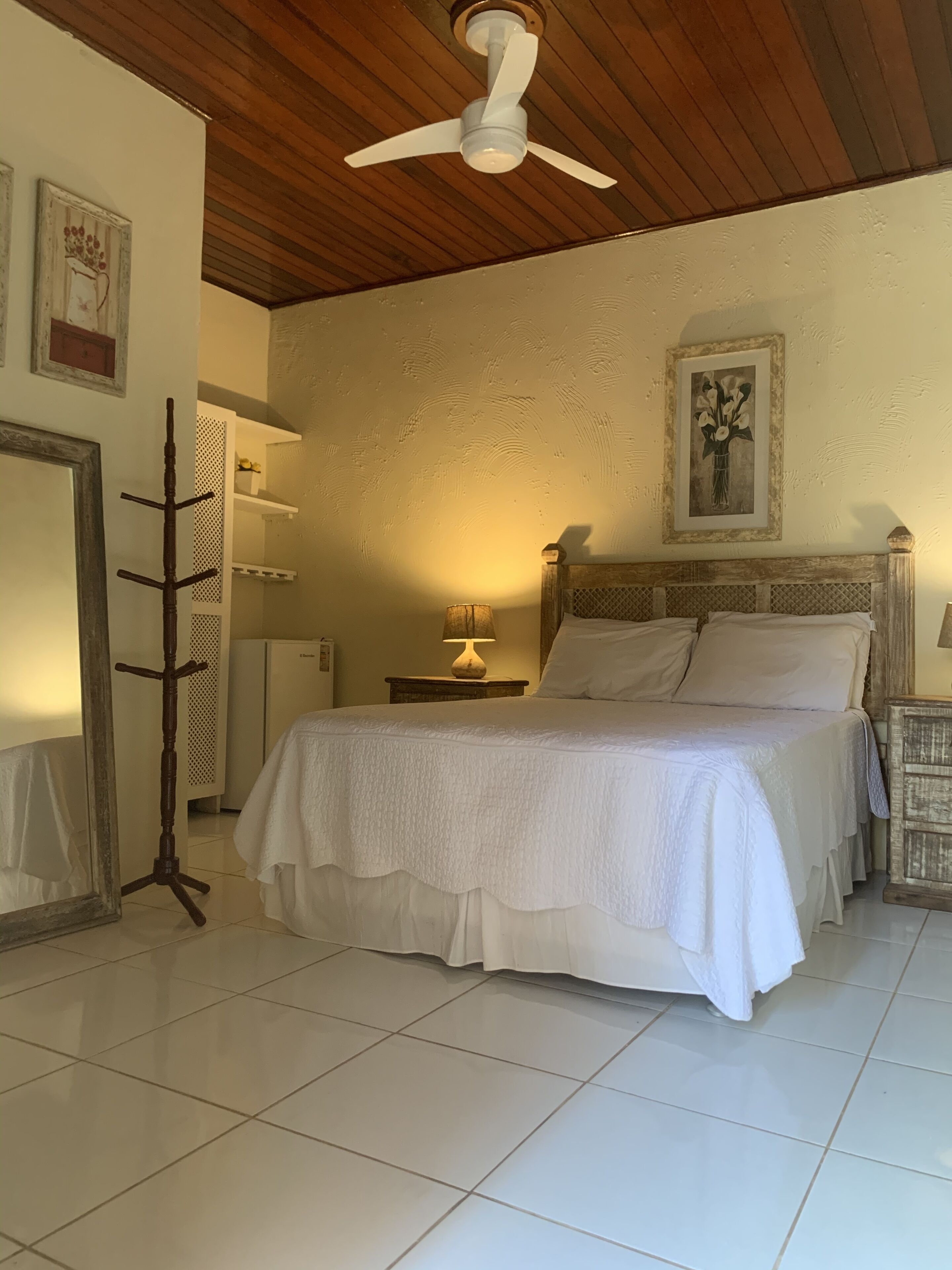 Quarto com cama, ventoinha de madeira no teto, mesa de cabeceira com abajur e um porta-roupas.
