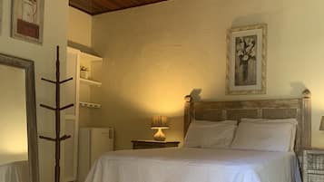 Quarto com cama, ventoinha de madeira no teto, mesa de cabeceira com abajur e um porta-roupas.