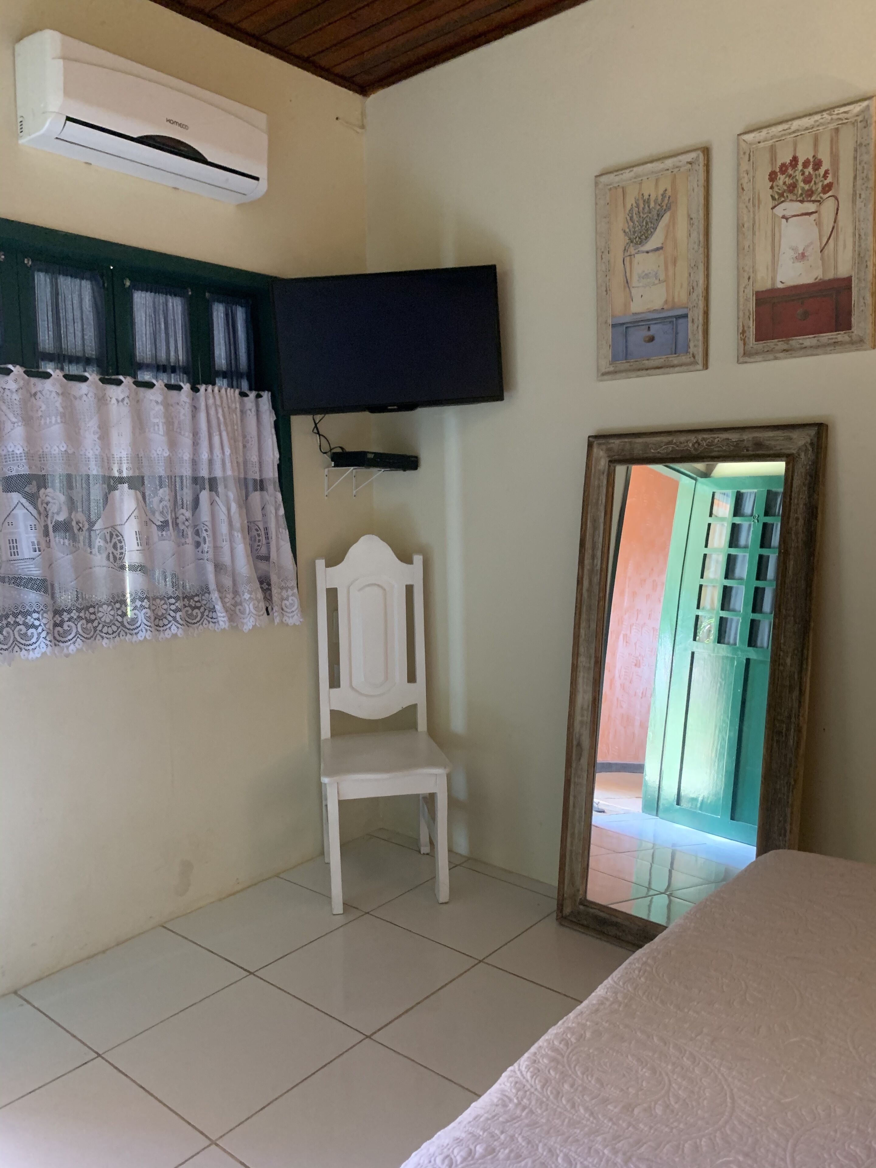 A room with a bed, a white chair, a TV mounted on the wall, and a window with curtains.