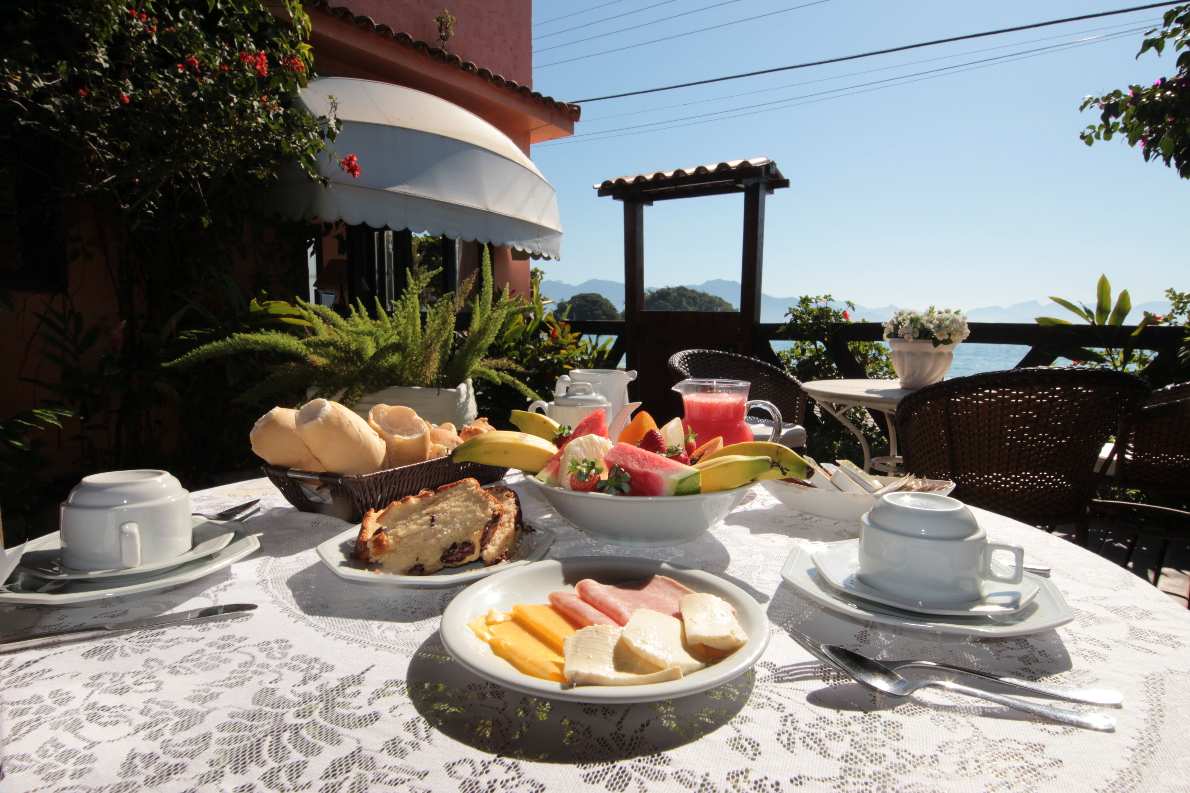 A table set for breakfast with various foods, including bread, fruits, and eggs, on a terrace with a view of the sea and mountains.